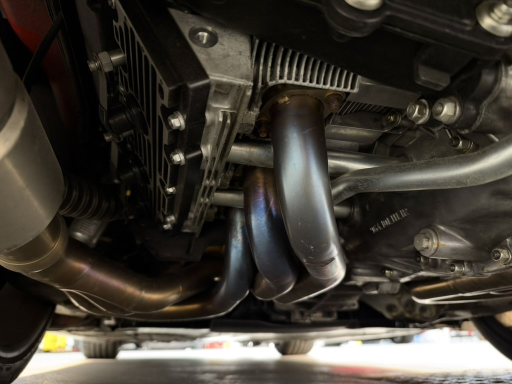 Underside view: M&M stainless long-tube headers merging under the engine, heat-coloured, with the oil cooler and tank above